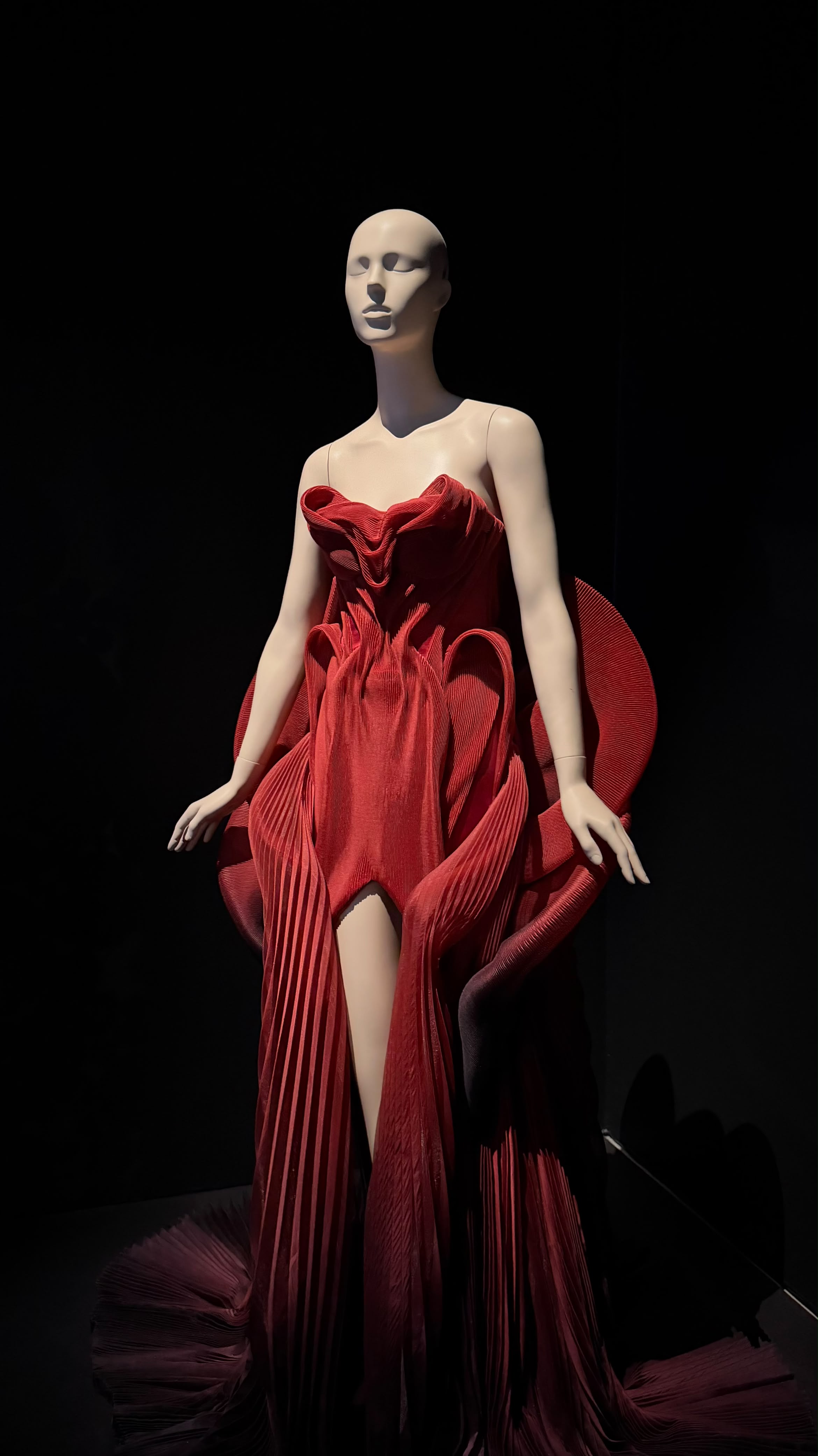 A red Iris van Herpen dress with layered sculptural folds against a dark exhibition wall