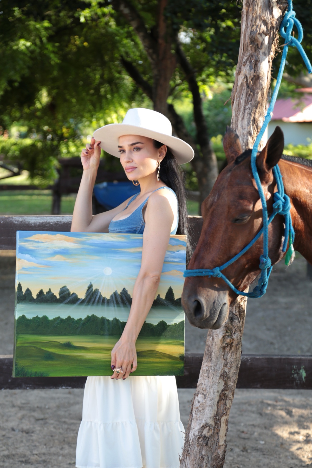 Galina in a white hat beside a horse, holding her green meadow painting
